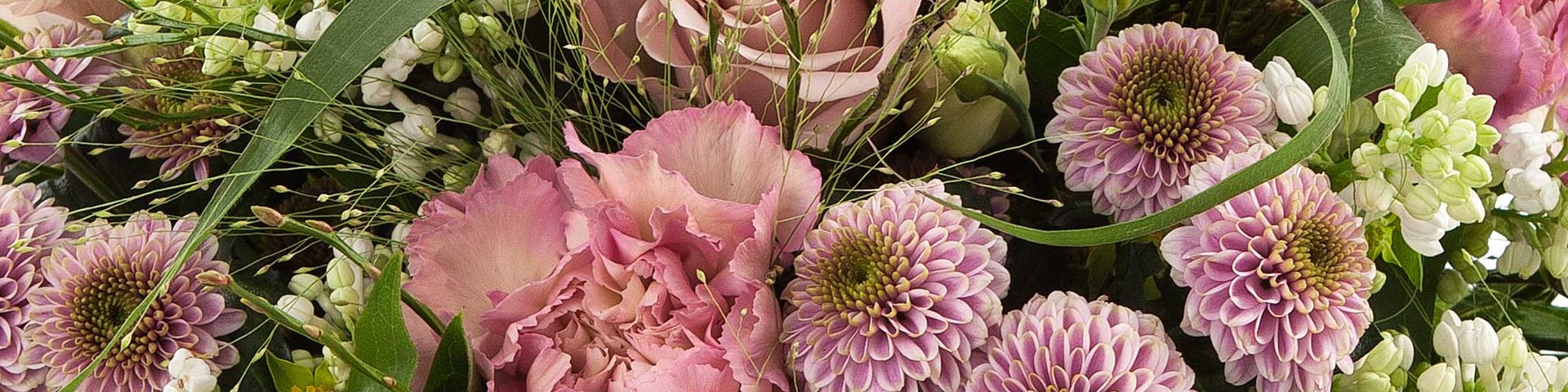 Et farverigt blomsterarrangement med lyserøde roser, dahliaer og nelliker. Arrangementet indeholder også grønne blade og mindre hvide blomster, hvilket skaber et frodigt og livligt udtryk.